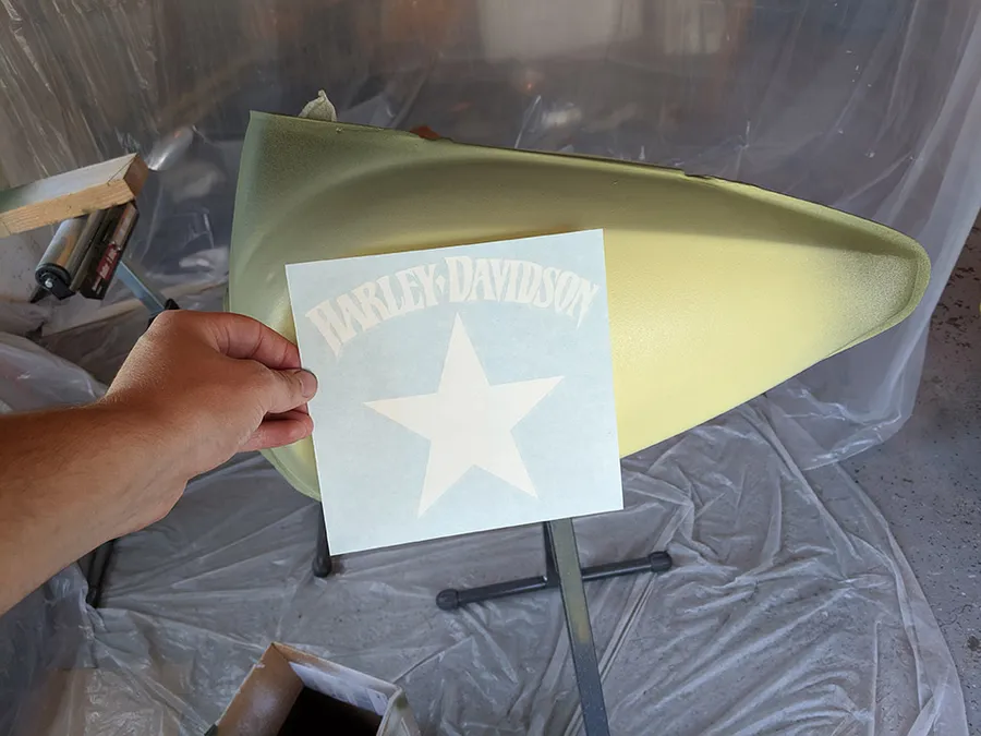 Hand holding a Harley-Davidson text and star positive stencil next to a motorcycle gas tank in pale yellow base coat